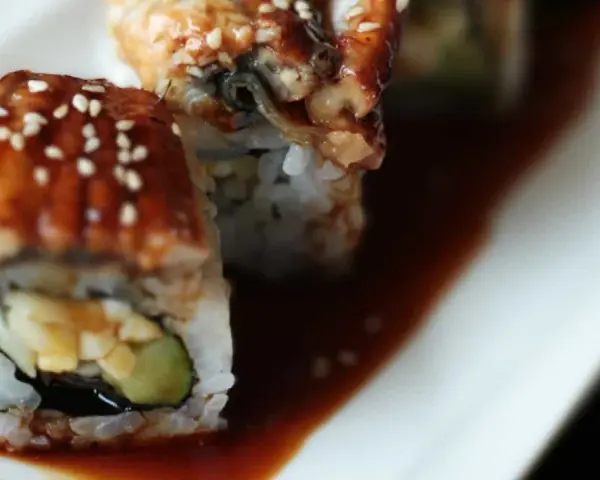 Rolka sushi z wężem morskim, polana słodkim sosem unagi i posypana sezamem, leży na talerzu.