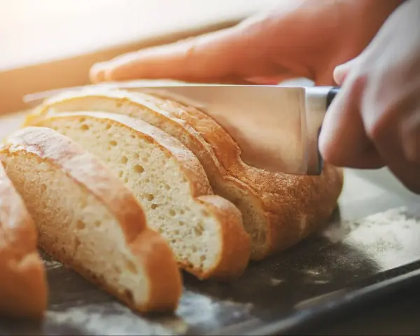 Dłoń kroi świeży chleb na kromki. Zastanawiasz się, jak odświeżyć chleb? Ten obrazek pokazuje, jak zacząć.