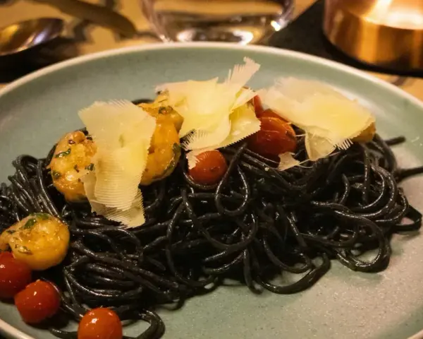 Czarny makaron z krewetkami, pomidorkami koktajlowymi i płatkami parmezanu na talerzu.