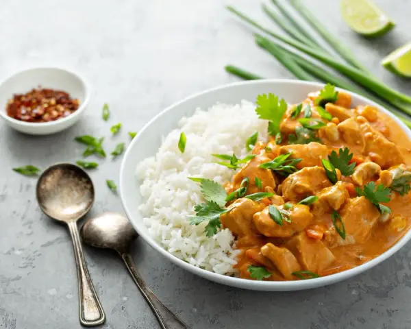 Pyszne curry masala z kurczakiem i ryżem, ozdobione szczypiorkiem i kolendrą. Obok miseczka z płatkami chili i ćwiartki limonki.
