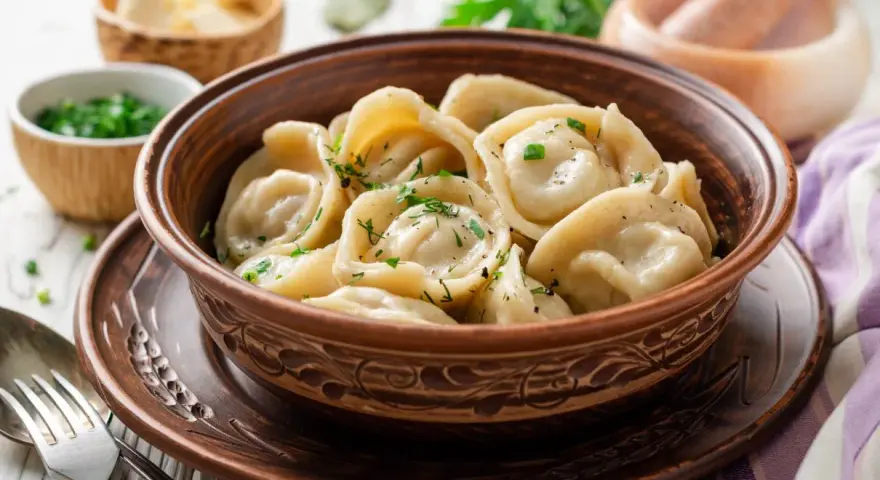 Pielmieni co to? Aromatyczne, gorące pierożki w glinianej misie, posypane zieloną natką. Obok miseczki z masłem i posiekanym szczypiorkiem.