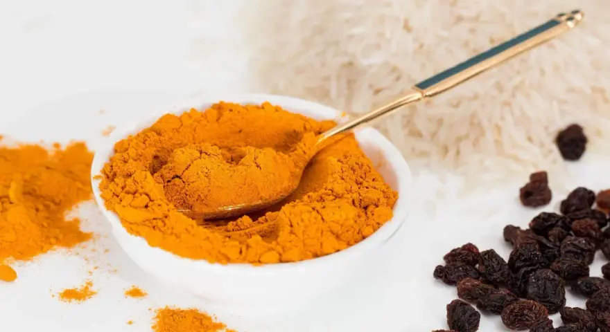 Miseczka pełna intensywnej przyprawy curry, obok ryż i rodzynki. Złota łyżeczka zanurzona w aromatycznym proszku.