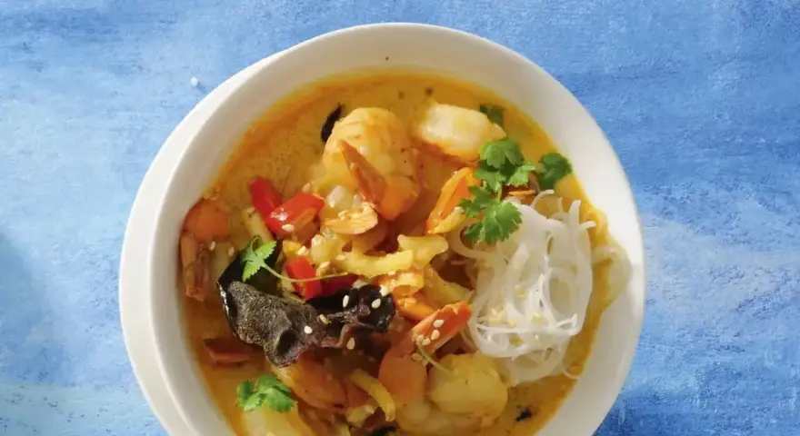Aromatyczna zupa krewetkowa z krewetkami, makaronem ryżowym, papryczką chili i kolendrą, podana w białej misce na niebieskim tle.