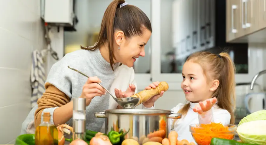 Mama i córka gotują pyszne zupy dla dzieci. Na stole warzywa: marchewka, ziemniaki, cebula, papryka i kapusta.