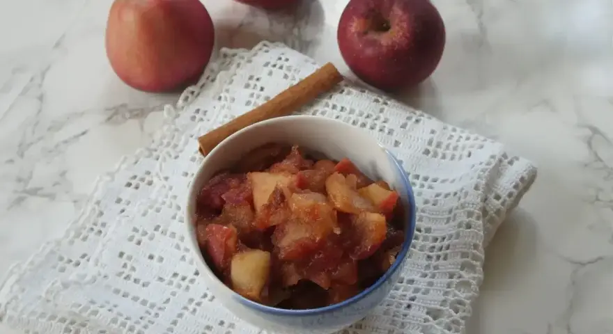 Miseczka z gotowanymi jabłkami do szarlotki, obok laska cynamonu i świeże jabłka.