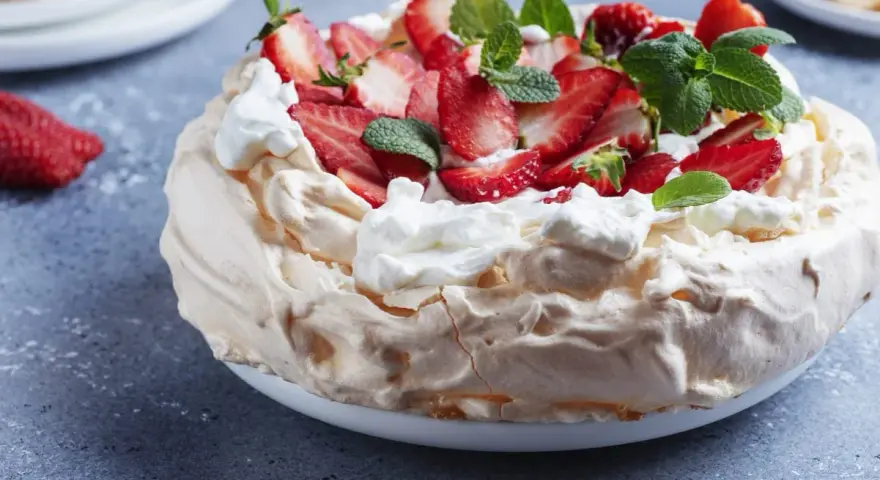 Puszysty tort bezowy z mascarpone i bitą śmietaną, ozdobiony świeżymi truskawkami i miętą.