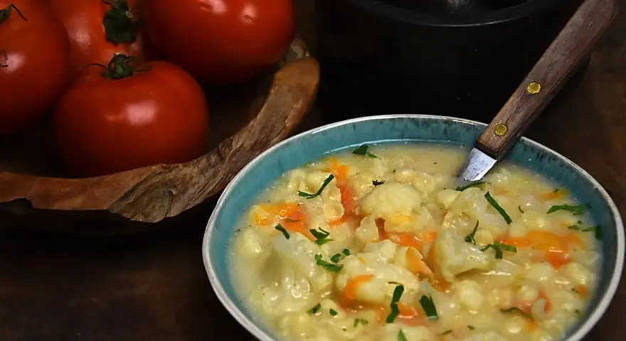 Aksamitna zupa kalafiorowa z makaronem, ozdobiona natką pietruszki. Obok miski leżą świeże pomidory.