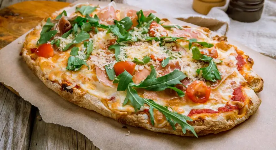 Pinsa co to? Podłużna, chrupiąca pizza z rukolą, pomidorkami i szynką parmeńską, posypana parmezanem.