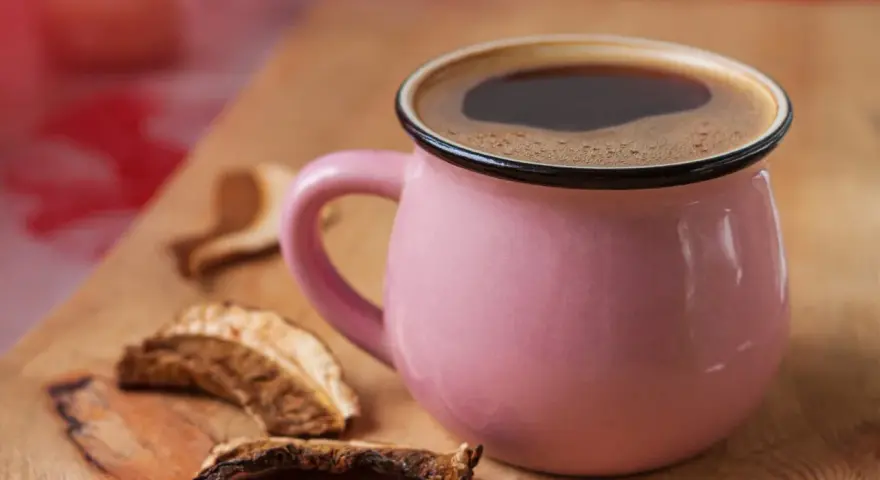 Różowy kubek z aromatyczną kawą grzybową na drewnianej desce, obok suszone grzyby.