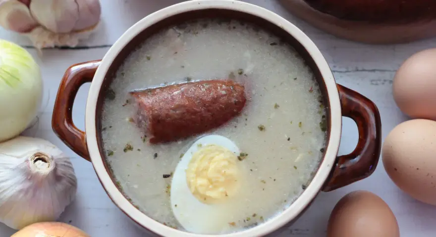 Aromaticzny żurek z kiełbasą i jajkiem, przygotowany na bazie domowego zakwasu na żurek. Obok czosnek, cebula i jajka.