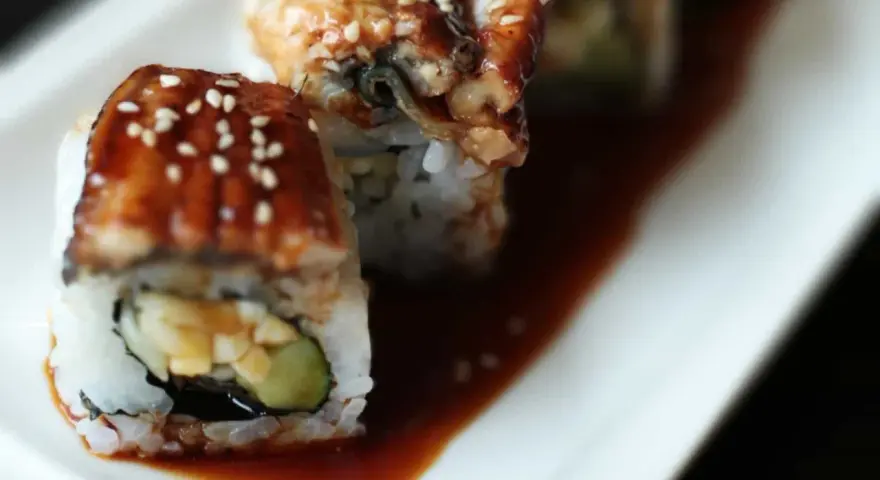 Rolka sushi z wężem morskim, polana słodkim sosem unagi i posypana sezamem, leży na talerzu.