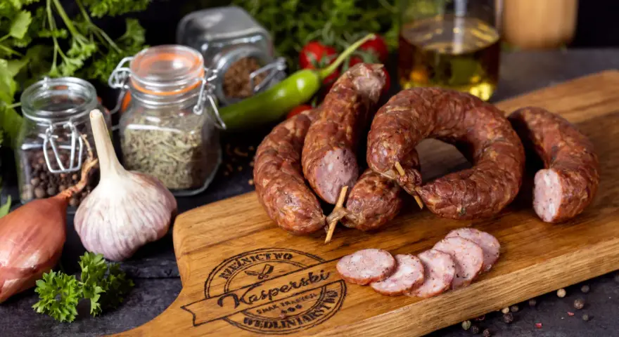 Na drewnianej desce leży pyszna, wędzona kiełbasa jałowcowa, obok czosnek, cebula i przyprawy.
