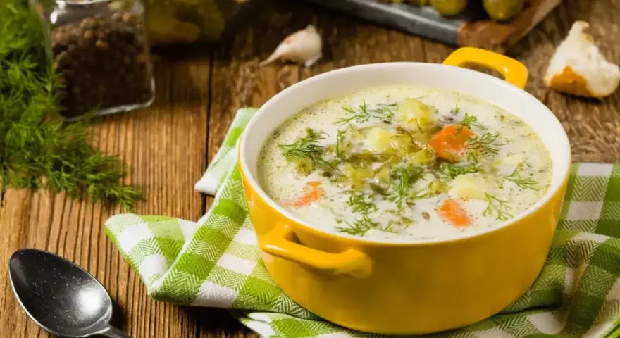 Zupa ogórkowa z koperkiem, ziemniakami i marchewką. W tle słoik z pieprzem, czosnek i ogórki kiszone. Idealne jakie przyprawy do zupy ogórkowej?