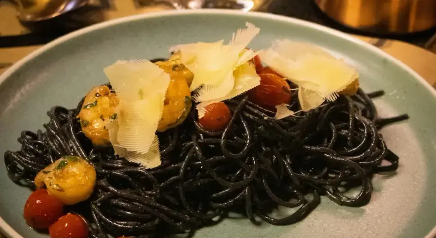 Czarny makaron z krewetkami, pomidorkami koktajlowymi i płatkami parmezanu na talerzu.