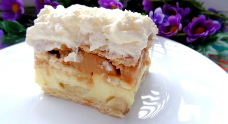 Szybkie ciasto z bananami i budyniem, warstwowe, z kremem i bitą śmietaną, na talerzu, w tle fioletowe kwiaty.