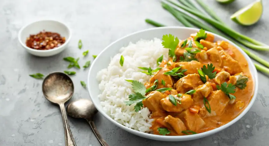 Pyszne curry masala z kurczakiem i ryżem, ozdobione szczypiorkiem i kolendrą. Obok miseczka z płatkami chili i ćwiartki limonki.
