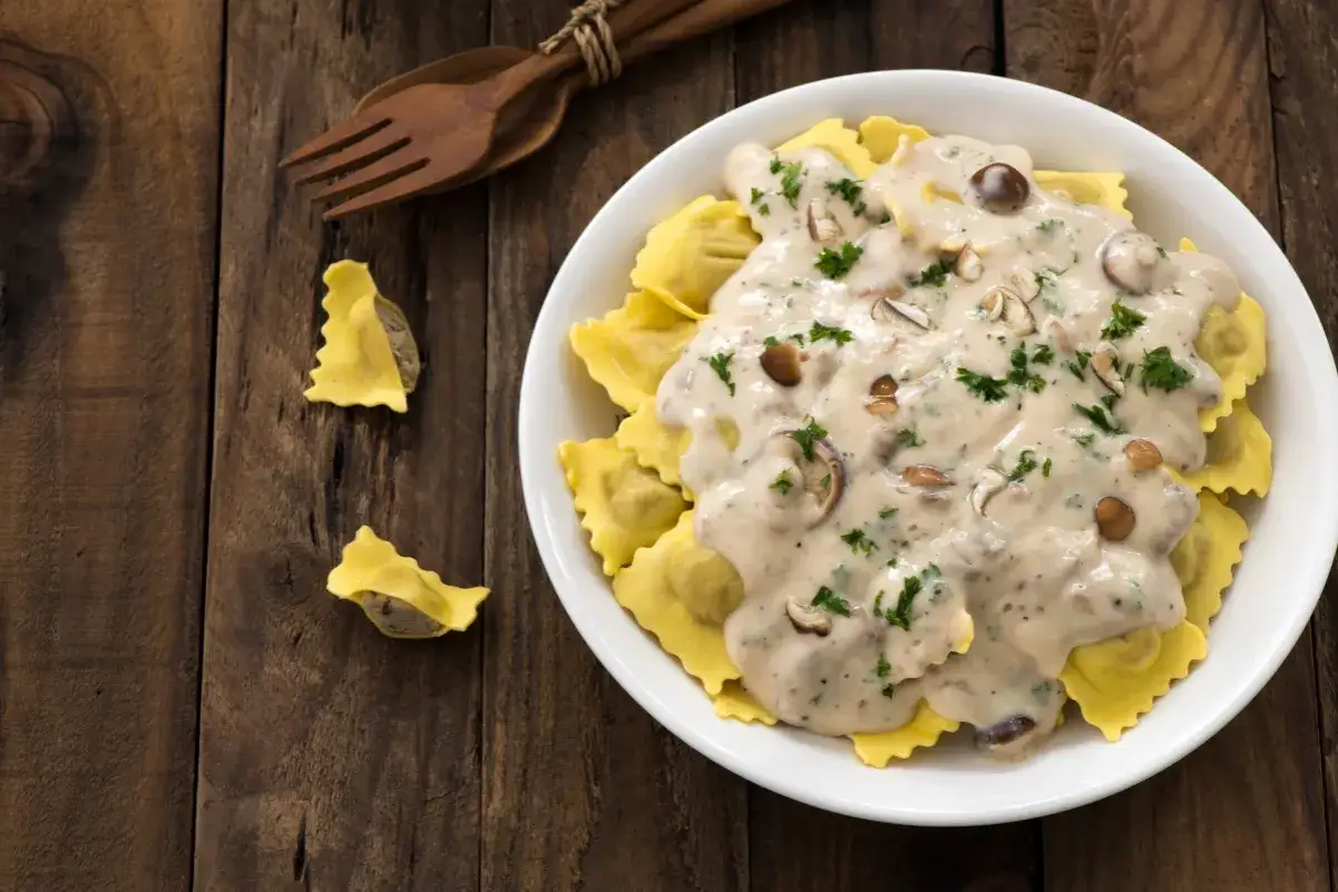 Ravioli w kremowym sosie śmietanowym z grzybami i natką pietruszki, podane na drewnianym stole.