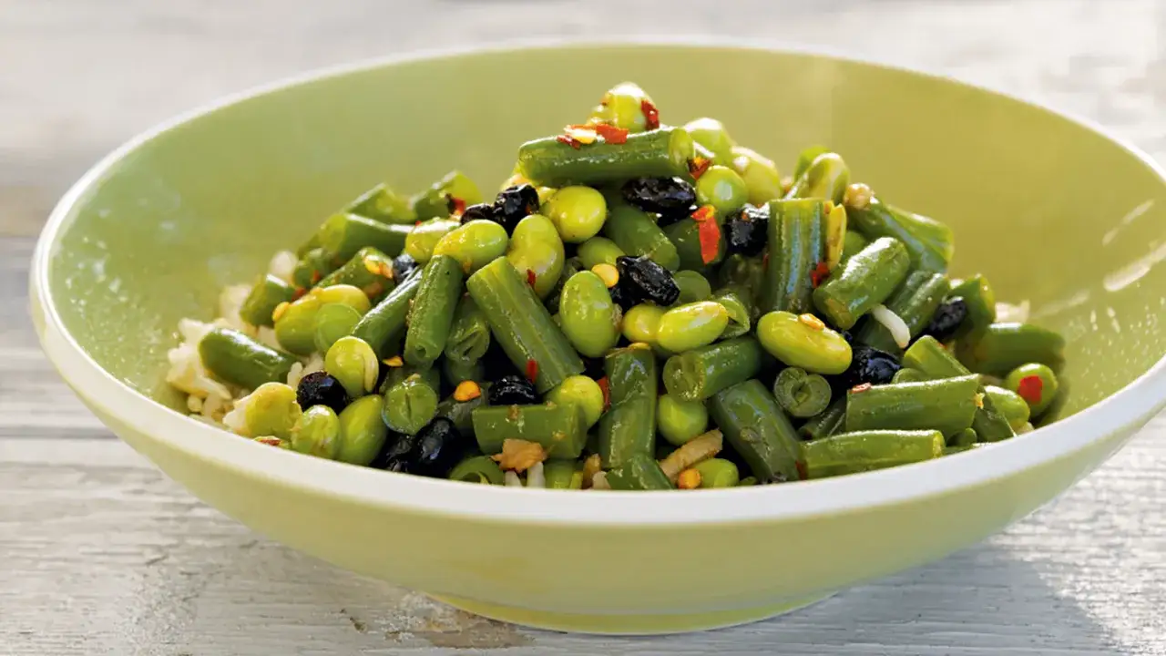 Miseczka pełna zielonych fasolek szparagowych i fasolki edamame, z dodatkiem czarnych oliwek i papryczki chili, podana na ryżu.