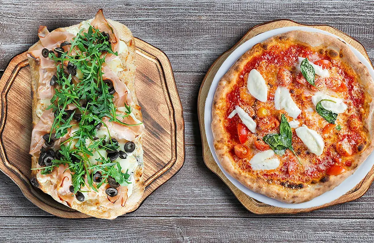 Pinsa co to? Dwie pyszne potrawy: prostokątna pinsa z szynką, rukolą i oliwkami oraz okrągła pizza Margherita z pomidorkami i mozzarellą.
