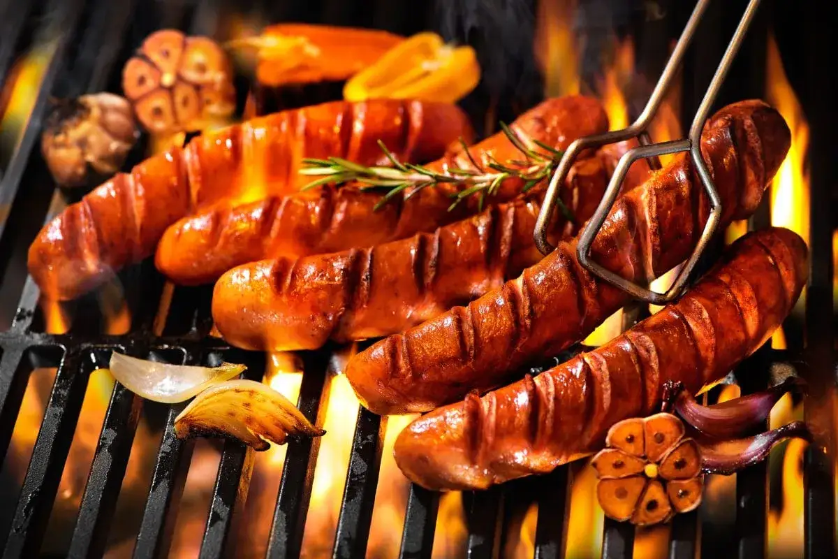 Soczysta kiełbasa na grilla skwierczy nad ogniem, obok pieczony czosnek i cebula. Idealna na letnie wieczory.