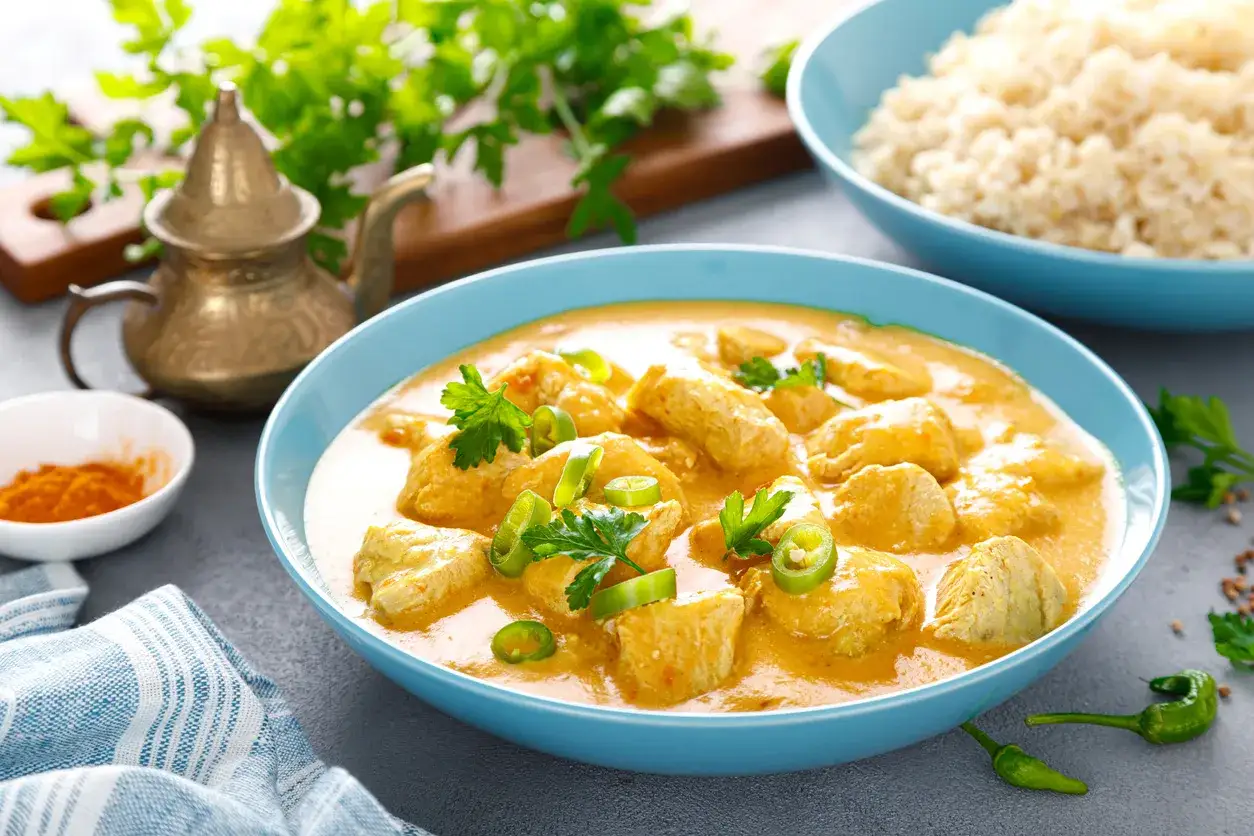 Aksamitny kurczak po tajlandzku w kremowym sosie, ozdobiony świeżą kolendrą i papryczkami chili. Obok miseczka ryżu i przypraw.