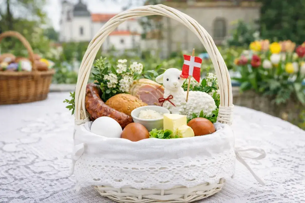 Biały koszyk z tradycyjnymi produktami do święconki: jajka, kiełbasa, chleb, masło, baranek z flagą.