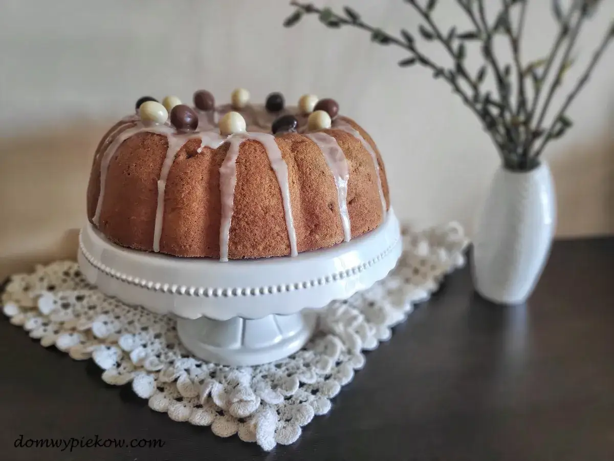 Wilgotna babka drożdżowa, ozdobiona lukrem i czekoladowymi jajeczkami, prezentuje się apetycznie na białej paterze.