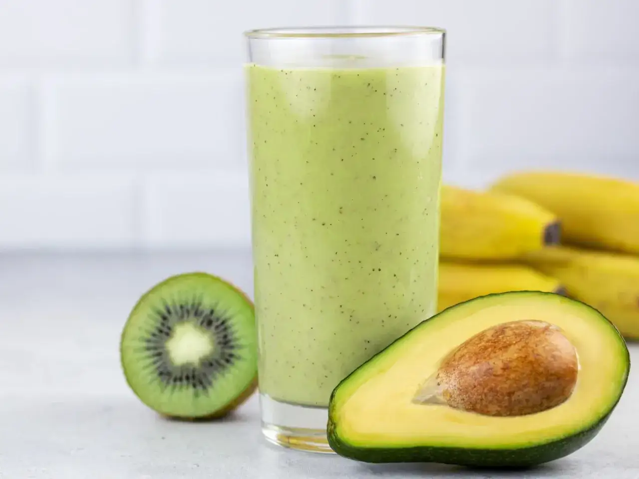 Zielony koktajl z awokado, kiwi i bananów w szklance, obok połówka awokado i przekrojone kiwi.