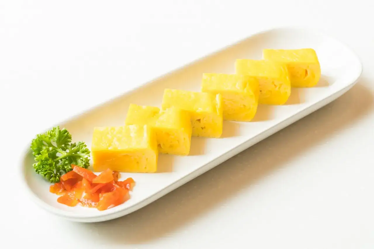 Sześć kawałków puszystego omletu tamago ułożonych na białej tacy, ozdobionej natką pietruszki i kawałkami pomarańczy.
