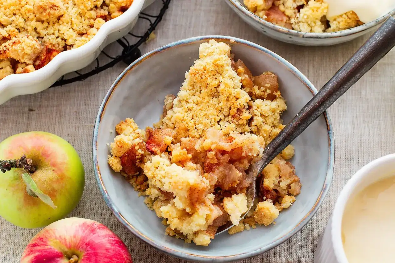 Szybkie ciasto na niedzielę do kawy: jabłkowy crumble z kruszonką, podany w miseczce z łyżką i jabłkami obok.