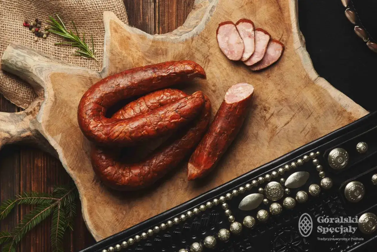 Na drewnianej desce leży pęta kiełbasy jałowcowej, obok kilka plastrów i kawałek. Całość ozdobiona jest góralskim pasem.